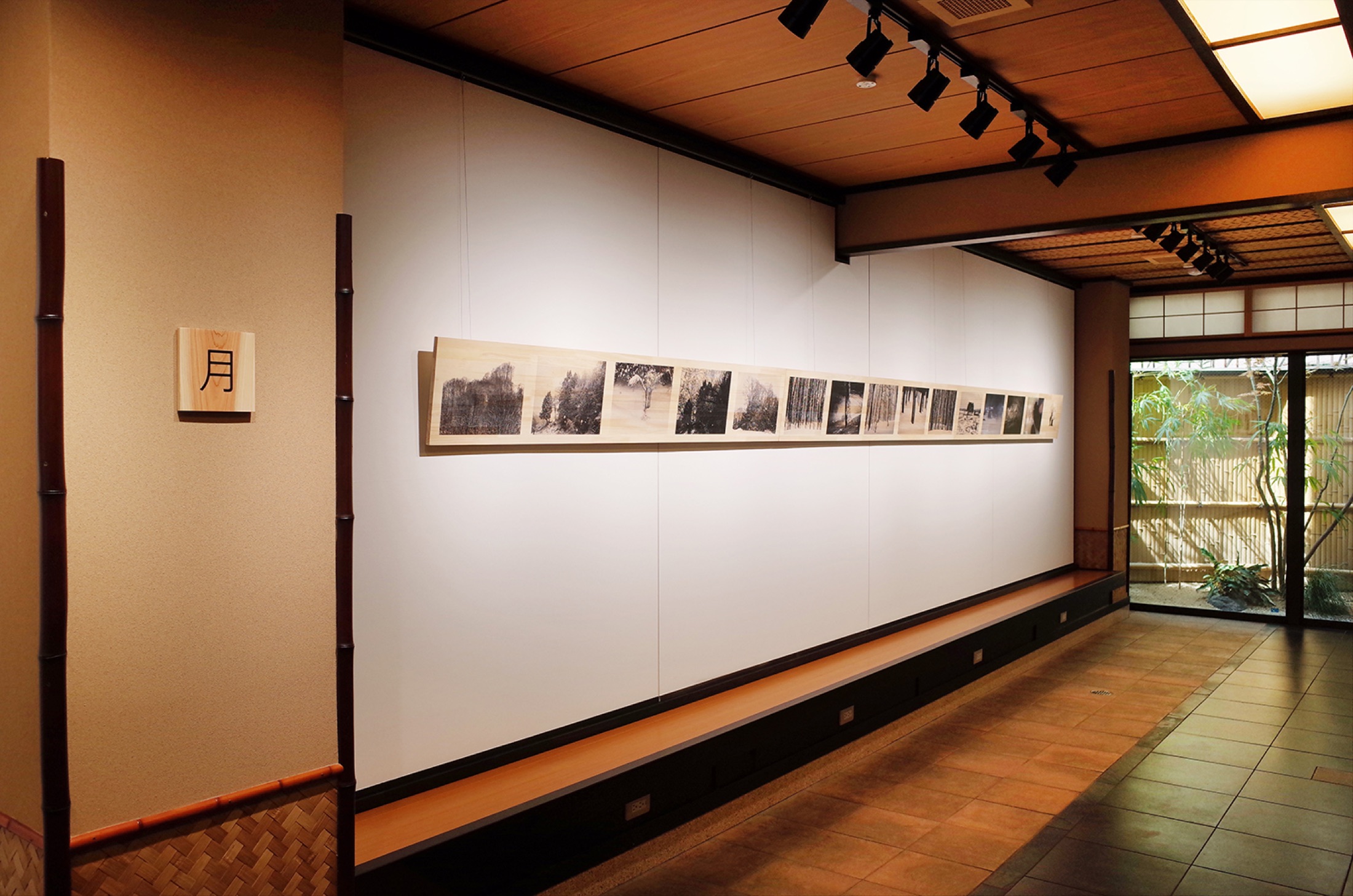 1F奥・庭側の展示室。白壁に作品が一列に並び、庭に向かって開く。柱には木札「月」